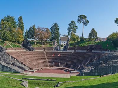 A wide-angle photograph of a large, semi-circular Roman theatre ruin with extensive stone seating (cavea) and a stage area, set within a grassy landscape with trees and modern houses in the background.