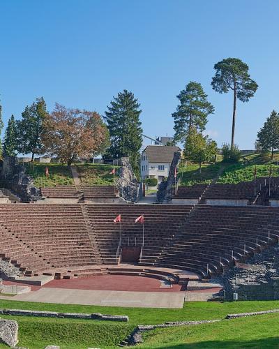 A wide-angle photograph of a large, semi-circular Roman theatre ruin with extensive stone seating (cavea) and a stage area, set within a grassy landscape with trees and modern houses in the background.