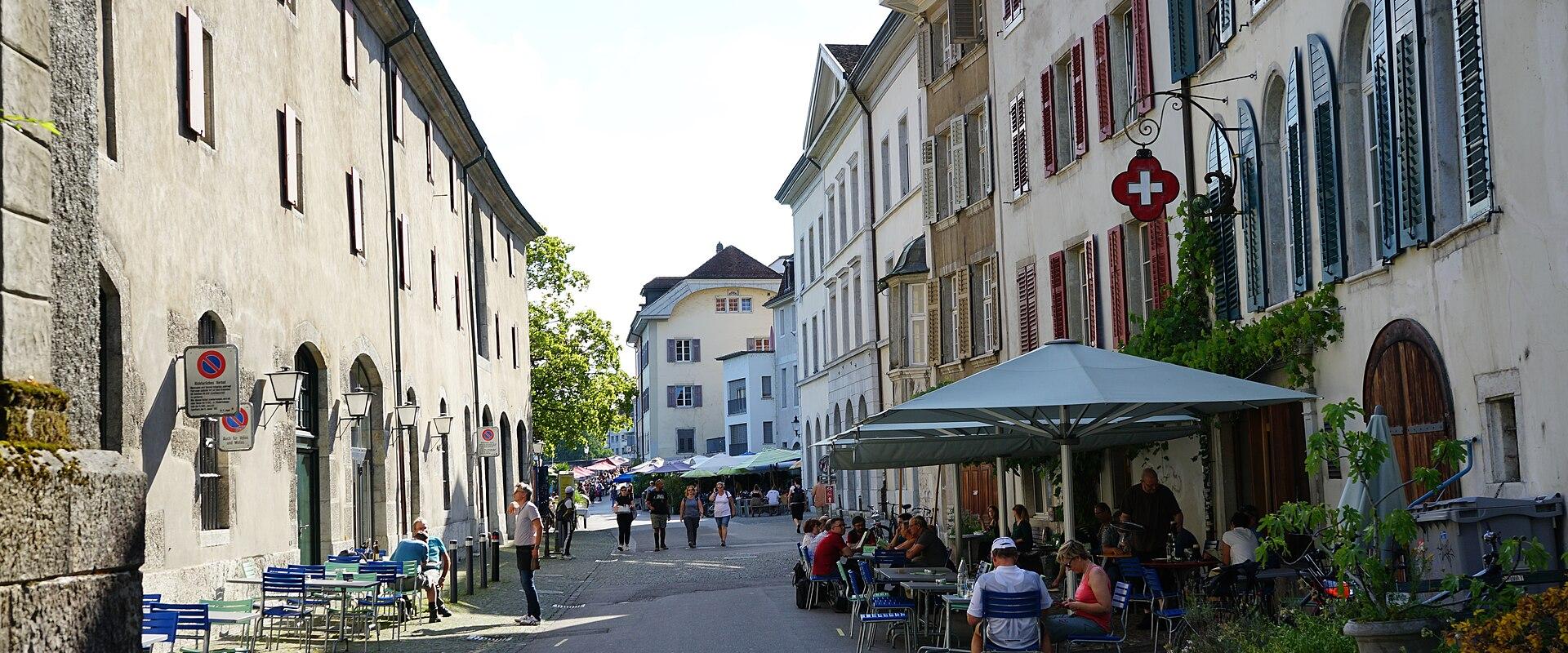 A narrow cobblestone street lined with historic buildings featuring shuttered windows and outdoor café seating, with a prominent Swiss flag sign hanging from a building on the right.