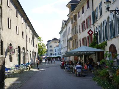 A narrow cobblestone street lined with historic buildings featuring shuttered windows and outdoor café seating, with a prominent Swiss flag sign hanging from a building on the right.