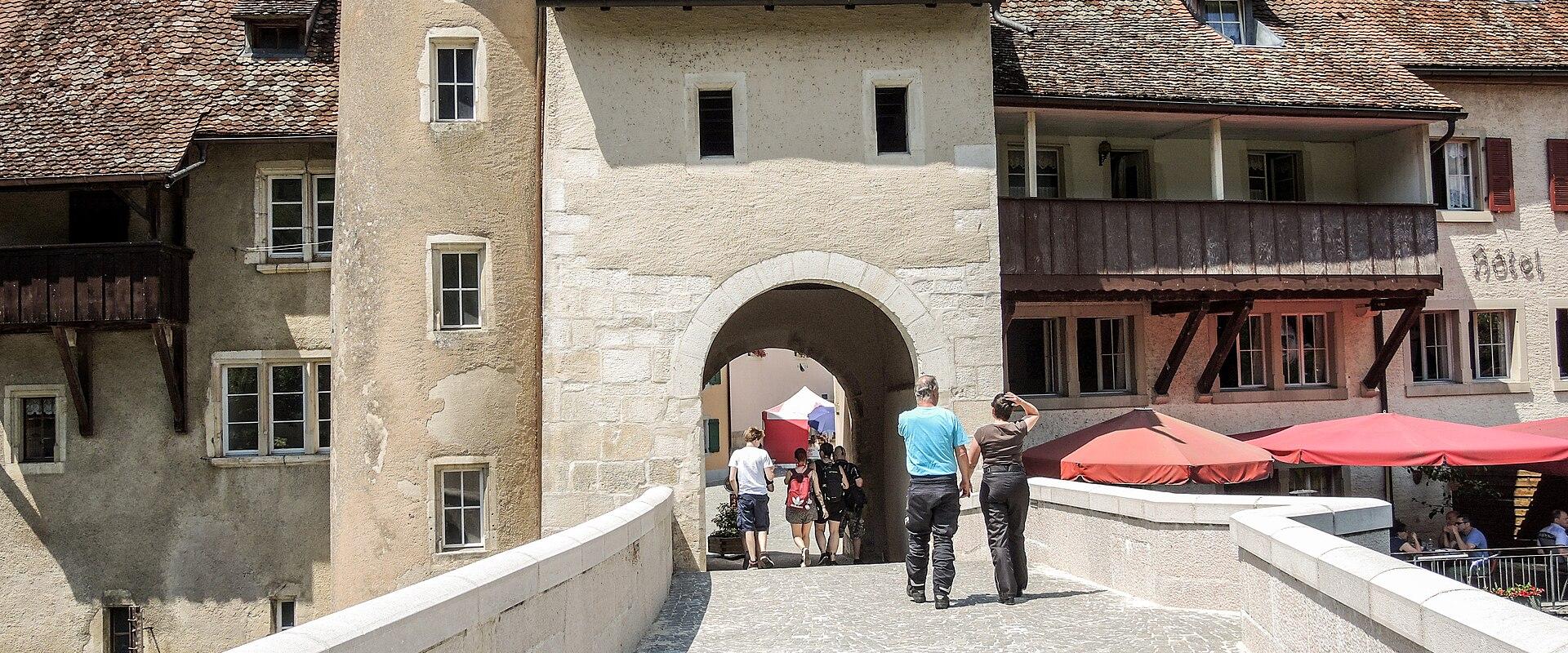 A stone bridge with a large arched gateway and a round tower on the left side, leading into a historic town square with red umbrellas and buildings.