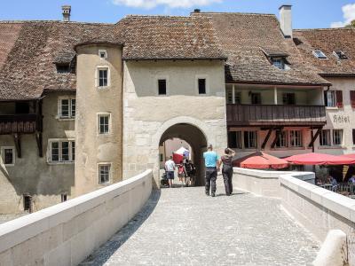 A stone bridge with a large arched gateway and a round tower on the left side, leading into a historic town square with red umbrellas and buildings.