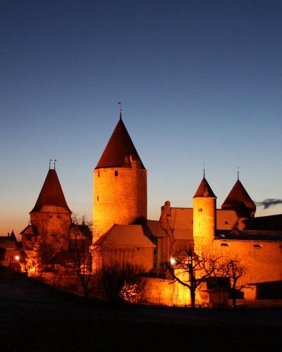 A medieval castle complex with multiple conical-roofed towers and stone walls illuminated at twilight, with a church spire visible in the background to the left.