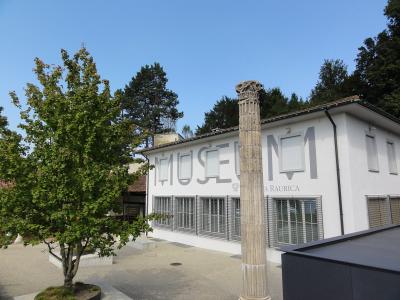 A white building with large grey lettering spelling "MUSEUM" and "A RAURICA" on its facade, with a single fluted stone column standing in the foreground.
