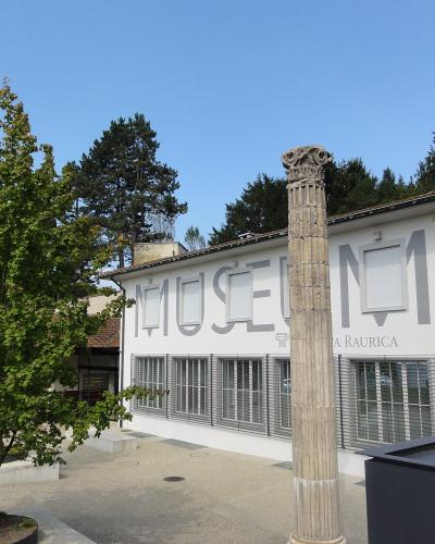 A white building with large grey lettering spelling "MUSEUM" and "A RAURICA" on its facade, with a single fluted stone column standing in the foreground.