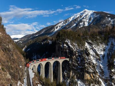 A red and white Glacier Express train crossing the arched stone Landwasser Viaduct in a snowy alpine valley.