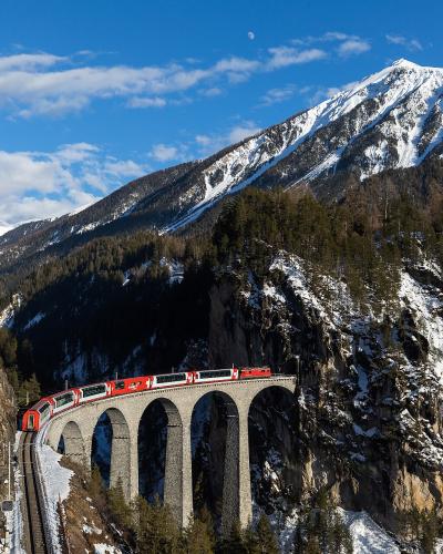 A red and white Glacier Express train crossing the arched stone Landwasser Viaduct in a snowy alpine valley.