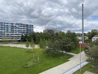 A modern urban park with green lawns, paved walkways, young trees, a tall metal mast, and multi-story residential buildings in the background under a cloudy sky.