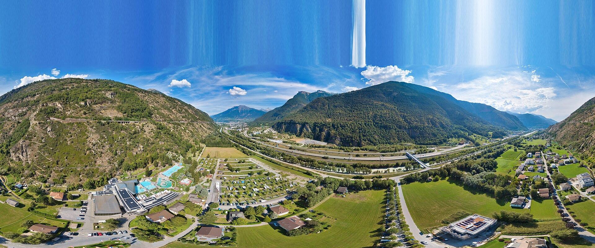 A panoramic aerial view of a valley in the Swiss Alps showing a large thermal spa complex with outdoor pools on the left, a campground, and a highway running through the center.