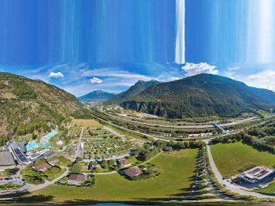 A panoramic aerial view of a valley in the Swiss Alps showing a large thermal spa complex with outdoor pools on the left, a campground, and a highway running through the center.