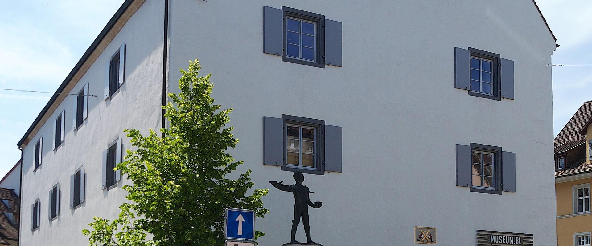 A large white building with a steep gable roof, featuring a statue of a figure on a fountain in the foreground and a sign reading "MUSEUM.BL" above an arched entrance.