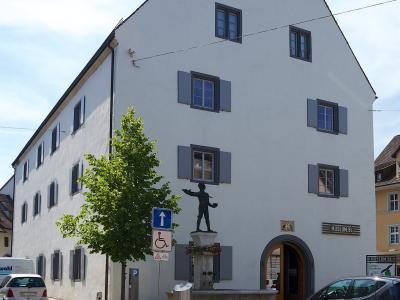 A large white building with a steep gable roof, featuring a statue of a figure on a fountain in the foreground and a sign reading "MUSEUM.BL" above an arched entrance.