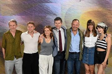 A group of seven people (cast and crew) posing for a photograph in front of a colorful, abstract backdrop.