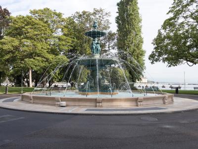 A large, ornate bronze fountain with multiple tiers and sculptural figures, situated in a paved circular plaza surrounded by green trees and a lake in the background.
