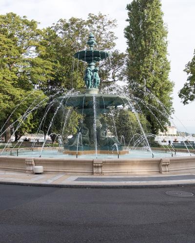 A large, ornate bronze fountain with multiple tiers and sculptural figures, situated in a paved circular plaza surrounded by green trees and a lake in the background.