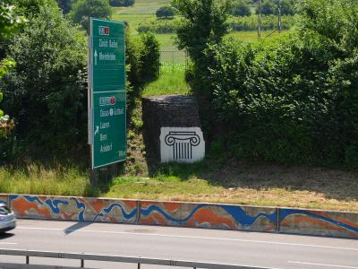 A green Swiss highway sign listing destinations like Zürich, Basel, and Arisdorf, standing next to a stone pillar featuring a painted Ionic column capital, with a painted concrete barrier in the foreground.