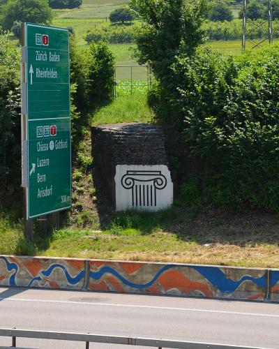 A green Swiss highway sign listing destinations like Zürich, Basel, and Arisdorf, standing next to a stone pillar featuring a painted Ionic column capital, with a painted concrete barrier in the foreground.