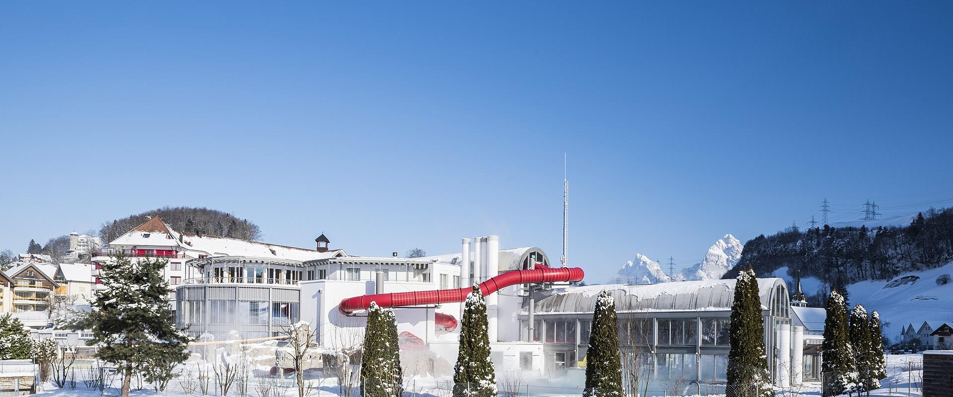 A snowy outdoor scene featuring a large white building complex with a distinctive long, red enclosed slide extending from it, set against a backdrop of snow-covered mountains and a clear blue sky.