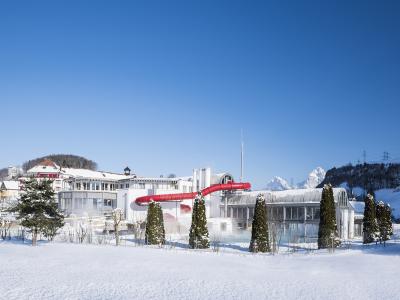 A snowy outdoor scene featuring a large white building complex with a distinctive long, red enclosed slide extending from it, set against a backdrop of snow-covered mountains and a clear blue sky.